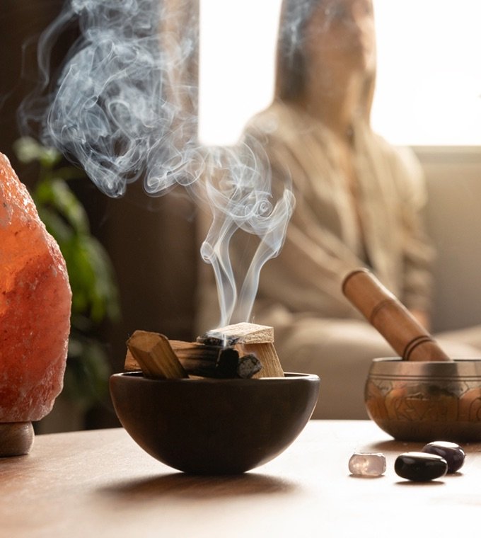 Incense smoke rising from a wooden bowl beside a singing bowl and salt lamp, with a person meditating in the background